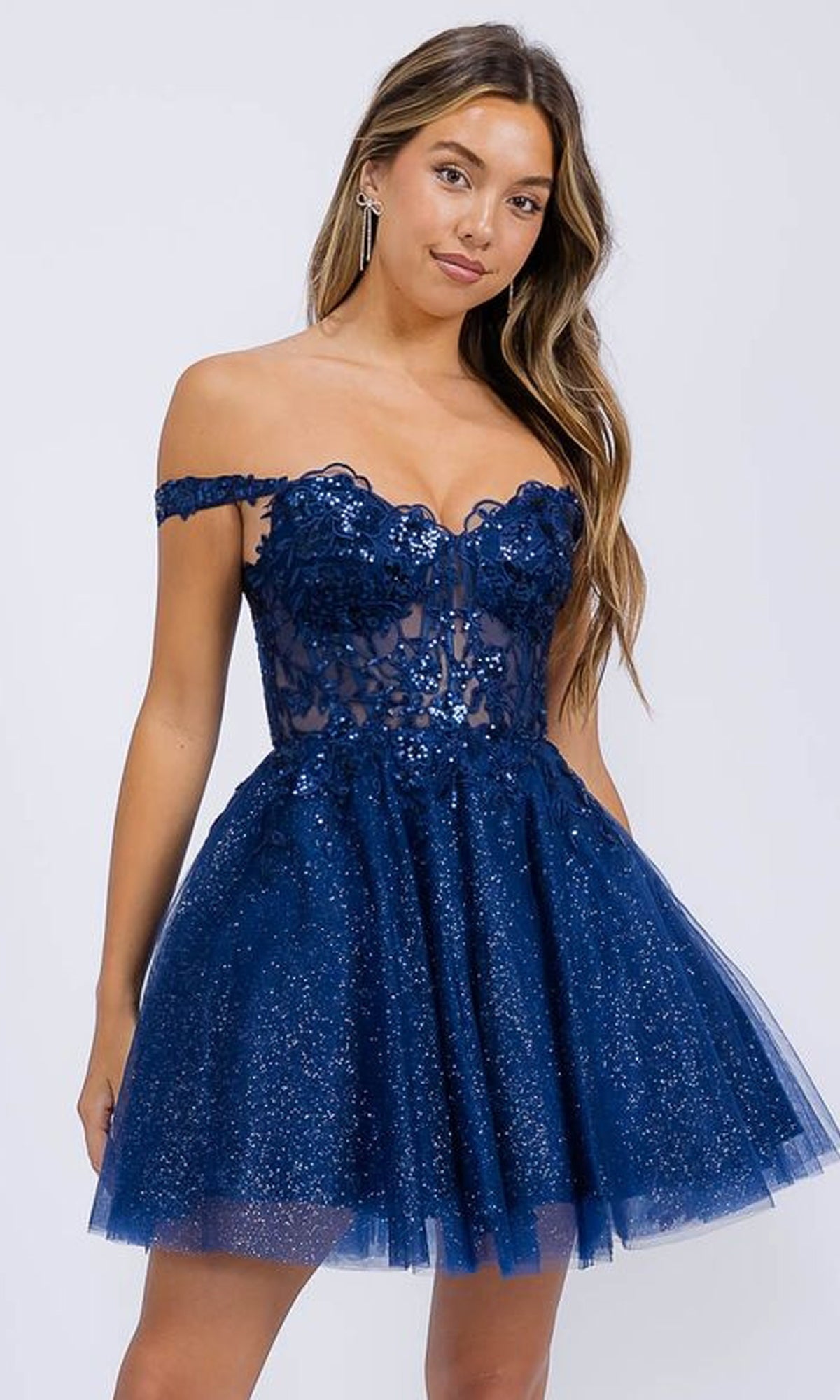 A woman models the Eureka EK138, a short navy blue off-the-shoulder homecoming dress with lace accents and a glitter tulle skirt, standing against a plain light background.